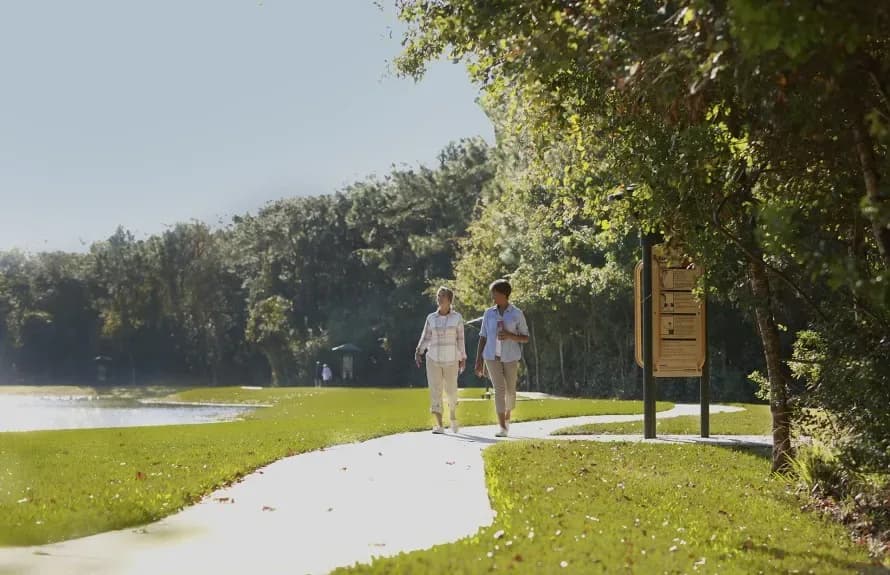 Two women walking on a paved trail beside a pond at the Del Webb Greenville community.
